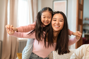 Cheerful Asian Mom Carrying Daughter Piggyback Having Fun At Home