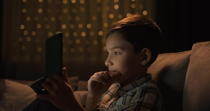 Adorable kid boy using digital tablet watching cartoons sitting on the sofa at night. Close up portrait of cute child watching cartoons video content relaxing at night time