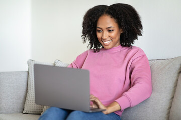 Cheerful millennial black woman typing on laptop, reads message, sitting on sofa in living room