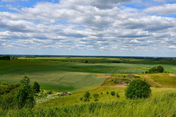 Fototapeta na wymiar A view from the top of a tall hill covered with grass, herbs, and other flora showing some vast fields, meadows, pasturelands, forest,moors, and a tall hill with some flags on top of it seen in summer