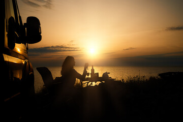 Outdoor leisure activity and wanderlust lifestyle: Silhouette of woman sitting at the camping table next to campervan against sunset, relax time. Lefkada Island, Greece.