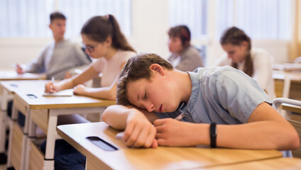 Tired teen student sleeping at desk in classroom during lesson with blurred classmates in background - Powered by Adobe