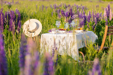 Romantic table decor for a loving couple on the blooming meadow with purple lupines. Two glasses of wine, flowers in a vase, silverware, fruits, wooden furniture and picnic basket. Sunset, golden hour