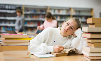 Bored teenager girl sitting at table in library while doing homework.