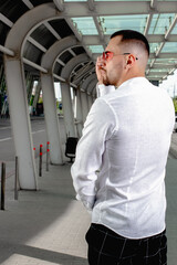 a handsome man nicely and stylishly dressed in pink glasses stands at a bus stop