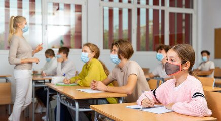 School girl in face mask sitting at desk in classroom on background with classmates and teacher