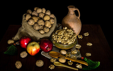 Sack of walnuts with and without shell, fresh apples and rustic utensils on a table and against a black background
