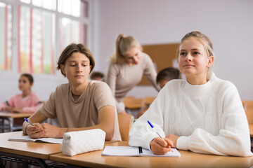 Teenage girl and boy sitting together at desk and doing tasks in classroom.