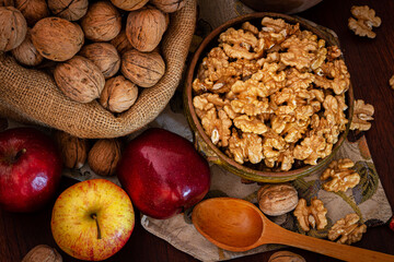 overhead view of a sack of walnuts with shell, clay plate with shelled walnuts,fresh apples, and a wooden spoon on a table