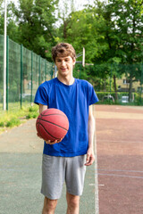 Cute boy in t shirt plays basketball on city playground. Active teen enjoying outdoor game with red ball. Hobby, active lifestyle, sport for kids.	