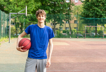 Cute boy in t shirt plays basketball on city playground. Active teen enjoying outdoor game with red...