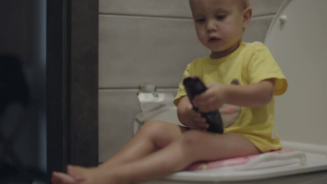 little baby child sit on toilet on pink child seat holding black TV remote hands. caucasian toddler boy yellow t-shirt learning use toilet. hygiene education preschooler health care concept. WC kid 