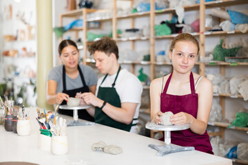 Young female teacher in apron helps teenager boy and teenager girl students to make product from clay in ceramic workshop