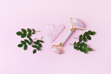 Modern selfcare concept. Pink jade roller and gua sha for face massage with green leaves. Facial massager tools. Anti age, lifting and toning care. Top view, flat lay.