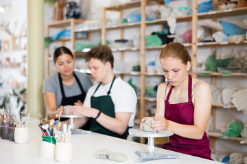 Young woman teacher shows students teenage girl and teenage boy how to sculpt ceramic product from raw clay in workshop