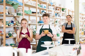 Teenage girl and guy show hand-made clay mugs. In the background, a satisfied female teacher