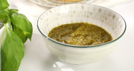 pesto sauce in bowl and on bread. basil and slices of bread on a white tablecloth. traditional italian green sauce. angle view. sunny day.