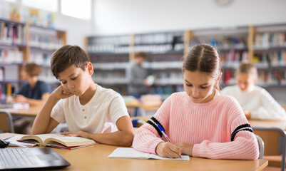 Focused ten-year-old schoolgirl, sitting at a desk in the library, writes a abstract in a exercise...