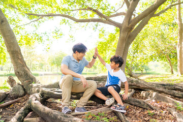 Happy Asian family enjoy and fun outdoor lifestyle on summer holiday vacation. Father and little son sitting under the tree and playing together at public park in the city. Family relationship concept