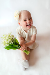 Close-up portrait of a smiling little girl in a white dress on white background. Toddler embraces a bouquet of fresh lilies of the valley . Gift for the holiday, the concept of purity, spring time. 