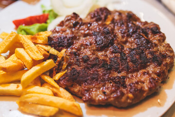 Pljeskavica with french fries on a plate, traditional serbian dish, grilled dish of spiced meat patty mixture of pork, beef and lamb, popular balkan steet food Pleskavitza, served in cafe - obrazy, fototapety, plakaty
