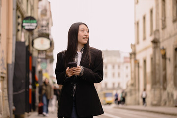 Young busy successful beautiful Asian business woman, korean professional businesswoman holding cellphone using smartphone standing or walking on old city street outside.