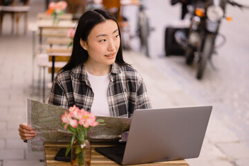 Portrait of a beautiful brunette Korean woman sitting in an authentic cafe and using a laptop, choosing travel routes on a map in an old European city.