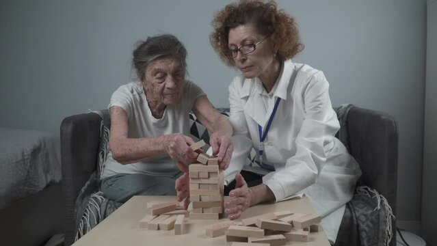 Senior woman practice skills build wooden blocks, building tower and trying not to let it fall, Jenga game. Old patient pull out block, place on top, support dotor during therapy dementia in house.