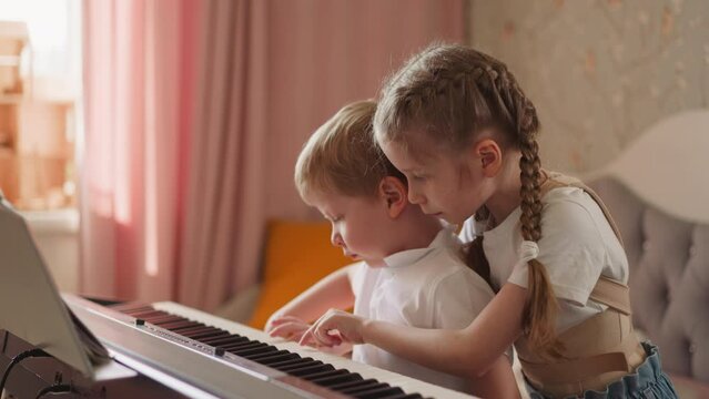Girl with orthopedic back braces plays piano hugging toddler blond boy in living room. Curious little brother looks at sister hands on instrument keyboard