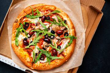 Hot pizza in box with spicy salami sausage, mozzarella cheese, mushrooms, black olives, tomato sauce and arugulal, just delivered, black table background, top view