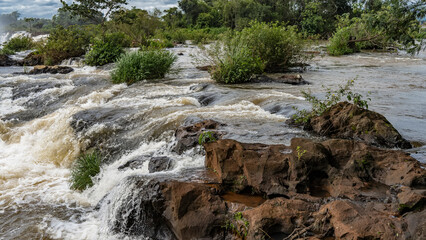 A stormy river rushes rapidly along a rocky bed. Foam and splashes. Boulders and islands of green vegetation in the water. Argentina. Iguazu.