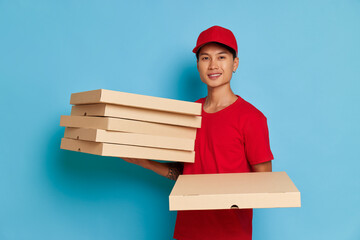 Cheerful Asian delivery guy holding some boxes with pizza in hands, wearing red cap and red t-shirt, fast delivery concept, copy space