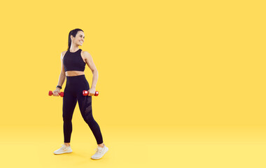 Fototapeta na wymiar Happy Caucasian woman with miniature dumbbells for fitness and performing physical activity looks back with smile in clothes for sports training stands on yellow studio background. Full length view