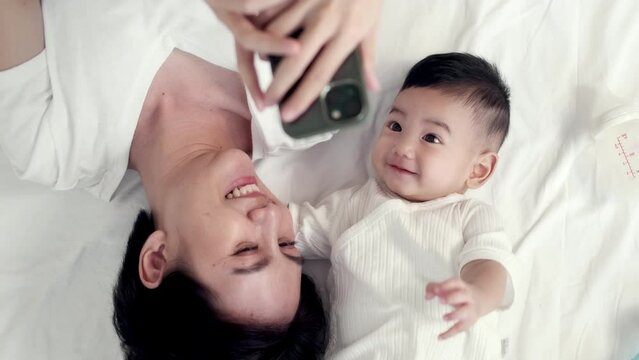Adorable asian baby and young mother joy making a selfie or video call to father or relatives in a bed. Concept of technology, new generation,family, connection, parenthood