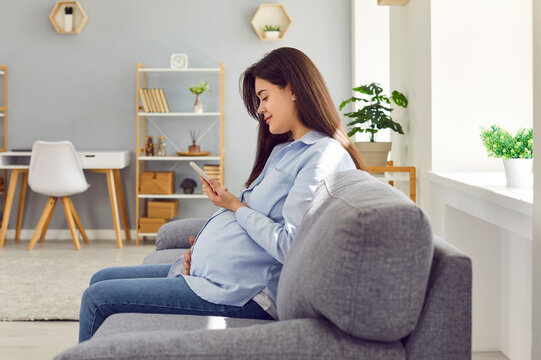 Side view of happy young pregnant woman sitting on sofa at home, holding her mobile phone, using pregnancy tracker app, looking at ultrasound picture of her baby or photos of children on social media