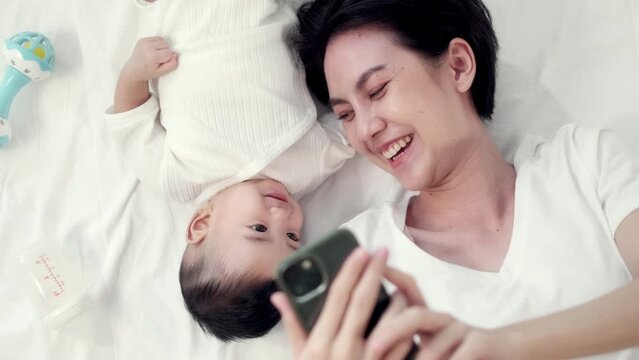 Happy Asian mother and her newborn baby making a selfie or video call to father or relatives in a bed. Concept of technology, new generation,family, connection, parenthood