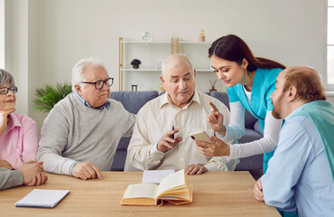 Obraz premium Young friendly female nurse helping senior people with mobile phone. Elderly men and women spending time together, reading a book in nursing home. Leisure in retirement home concept.