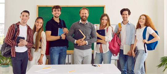 Photo of classmates and teacher standing against green chalk board on background. End of semester, happy, successful students are standing with professor holding textbooks in hands.