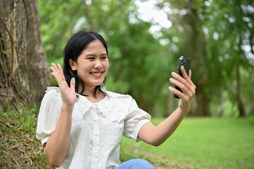 A cheerful Asian woman enjoys talking with her friend on a video call while relaxing in the park.