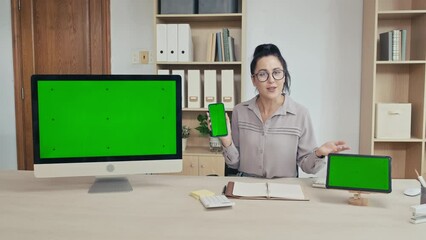 Medium shot of young woman presenting software on computer, smartphone, digital tablet with green chroma key screen looking at camera in office - Powered by Adobe