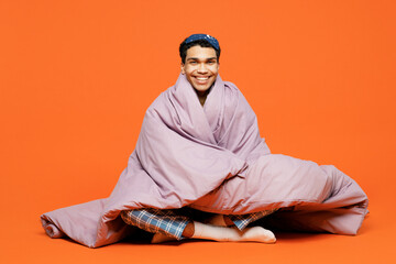 Full body smiling happy young man wear pyjamas jam sleep eye mask sit wrapped in blanket look camera rest relax at home isolated on plain orange background studio portrait. Good mood night nap concept
