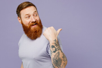 Young redhead bearded man wears violet t-shirt casual clothes point thumb finger aside indicate on...