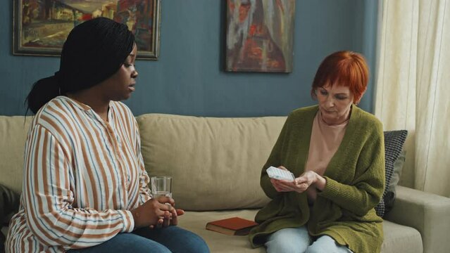 Medium shot of African American home care assistant giving glass of water to senior woman with red hair taking pills, both sitting on couch in living room