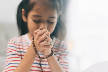 Asian little girl praying with rosary in hand at home.