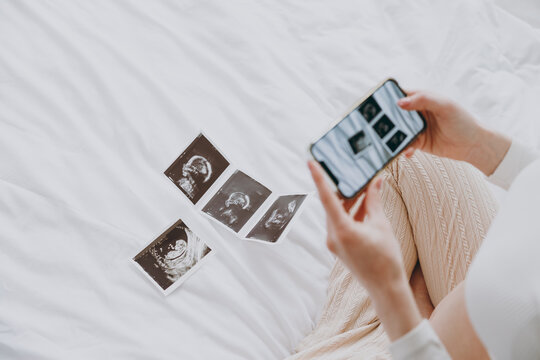 Cropped young pregnant woman wearing pyjamas taking photo of ultrasound examination on mobile cell phone rest relax spend time in bedroom lounge home. Maternity family pregnancy expectation concept.
