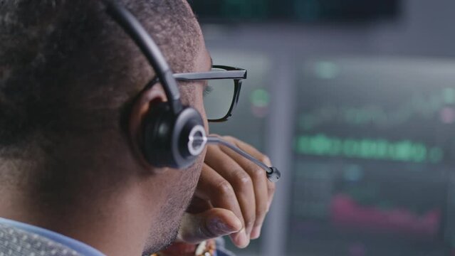 African American financial analyst in headset working in bank office. Businessman looks at computer screen, monitors real-time stocks, exchange market charts. Investment and analytics. Close up.