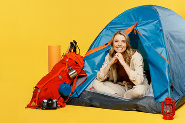 Full body smiling happy young woman sit near bag with stuff tent look camera isolated on plain yellow background. Tourist leads active lifestyle walk on spare time Hiking trek rest travel trip concept
