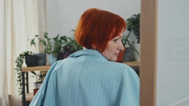 Medium closeup of cheerful senior woman trying on blue jacket in front of mirror in cozy studio at daytimeMedium closeup of cheerful senior woman trying on blue jacket in front of mirror in cozy studi