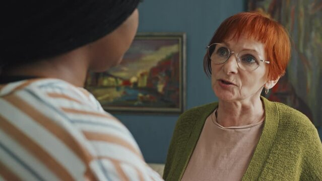 Over-the-shoulder shot of upset senior woman talking about her feeling to African American home care assistant in living room at daytime