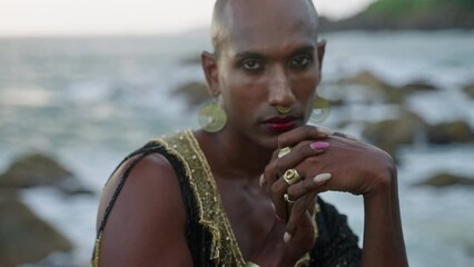 Queer black person in luxury dress, handmade jewelry poses on scenic sea shore. Closeup slow motion. Lgbtq biethnic fashion gay model in posh gown, brass accessories portrait. Tropical location.